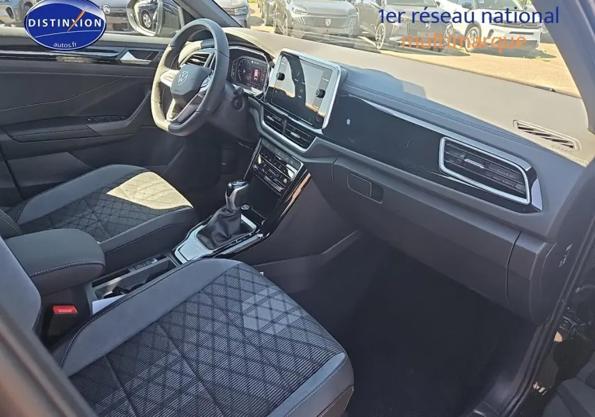 Intérieur du Volkswagen T-Roc 2025 vu côté passager, sièges tissu noir à motifs, tableau de bord moderne et écran tactile.