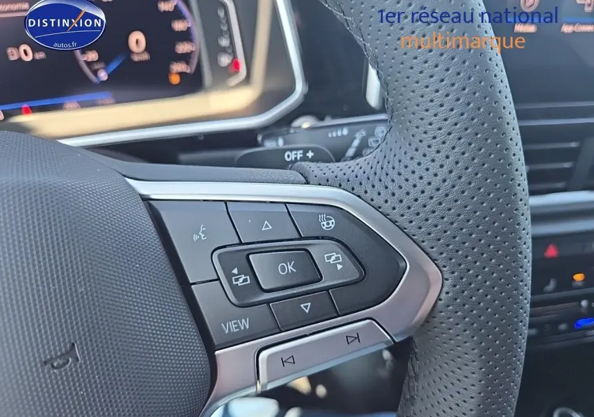 Gros plan sur le volant cuir perforé du Volkswagen T-Roc noir perle, avec commandes multifonctions et tableau de bord numérique flou.