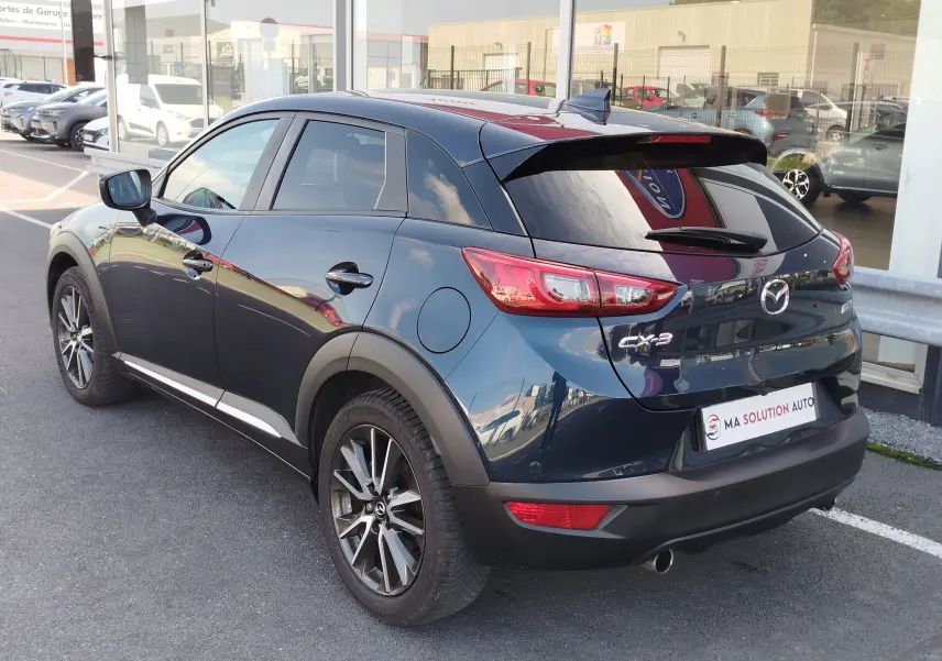 Vue 3/4 arrière droite du Mazda CX-3 bleu Dynamic Mica avec jantes alliage et vitres teintées, stationné devant un showroom.