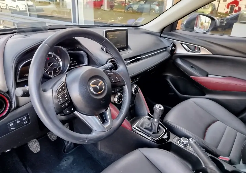 Intérieur du Mazda CX-3 2016 vu côté conducteur, volant cuir, sièges noirs avec surpiqûres rouges et levier de vitesse manuel.