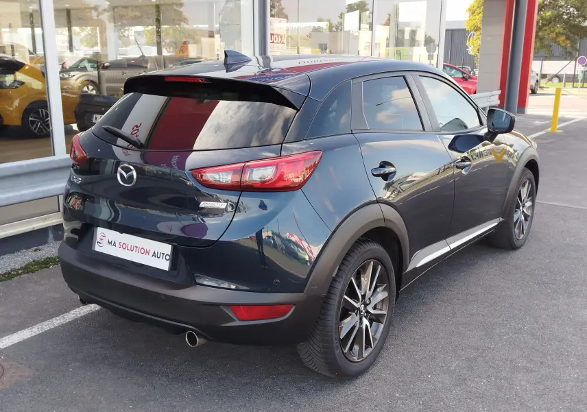 Vue 3/4 arrière droite d'un Mazda CX-3 bleu foncé avec jantes alliage et vitres teintées, stationné devant un showroom.