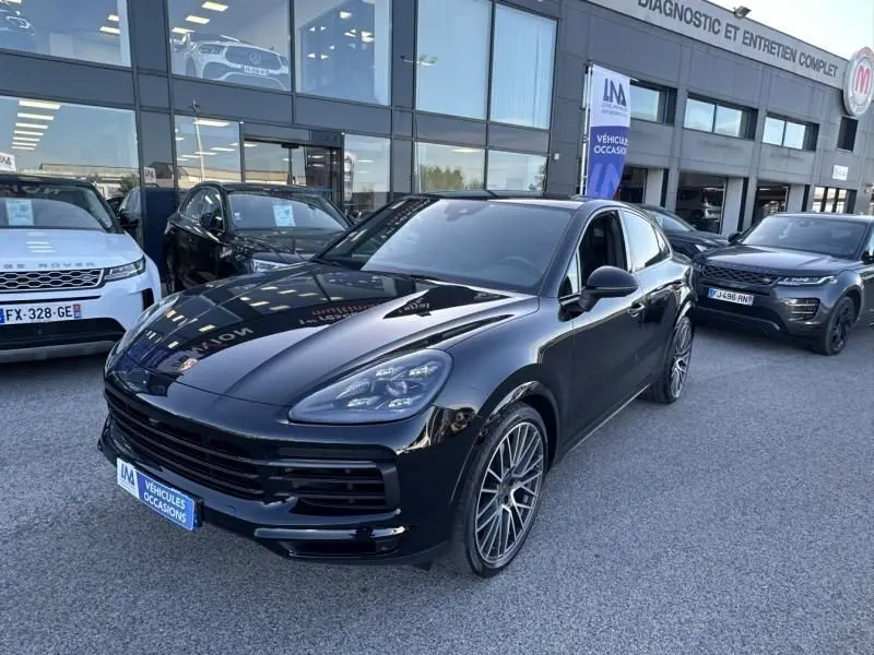 Vue 3/4 avant d'un Porsche Cayenne Coupé noir métal 2020, avec jantes alliage et calandre large, stationné devant un garage.