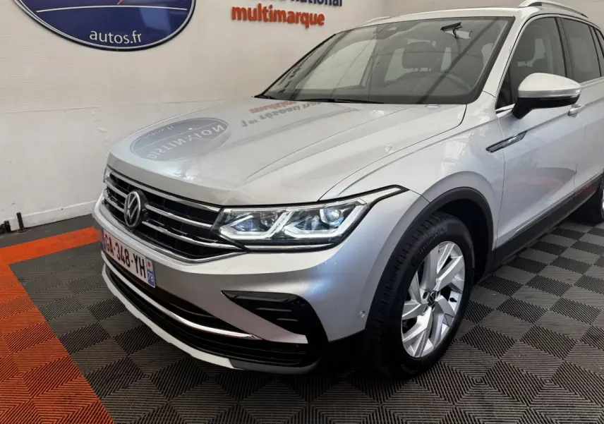 Vue 3/4 avant droit d’un Volkswagen Tiguan gris clair avec calandre chromée et phares LED allumés en intérieur showroom.