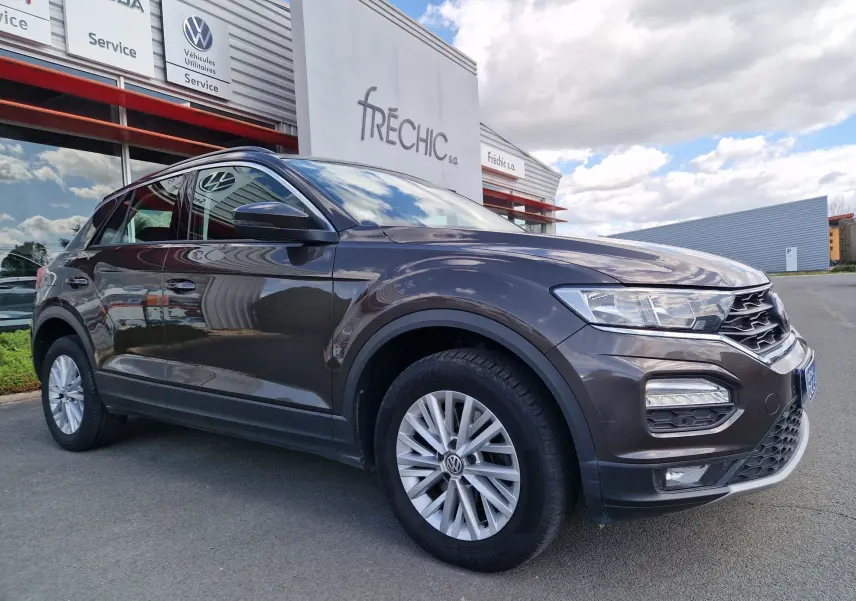 Volkswagen T-Roc chêne noir en 3/4 avant droit, avec jantes alliage et calandre distinctive sous un ciel nuageux.