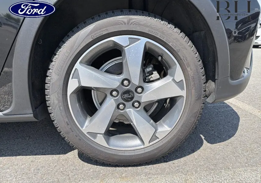 Gros plan sur la roue avant gauche d'une Ford Focus noire métallisée, jante alliage 5 branches avec logo Ford central.