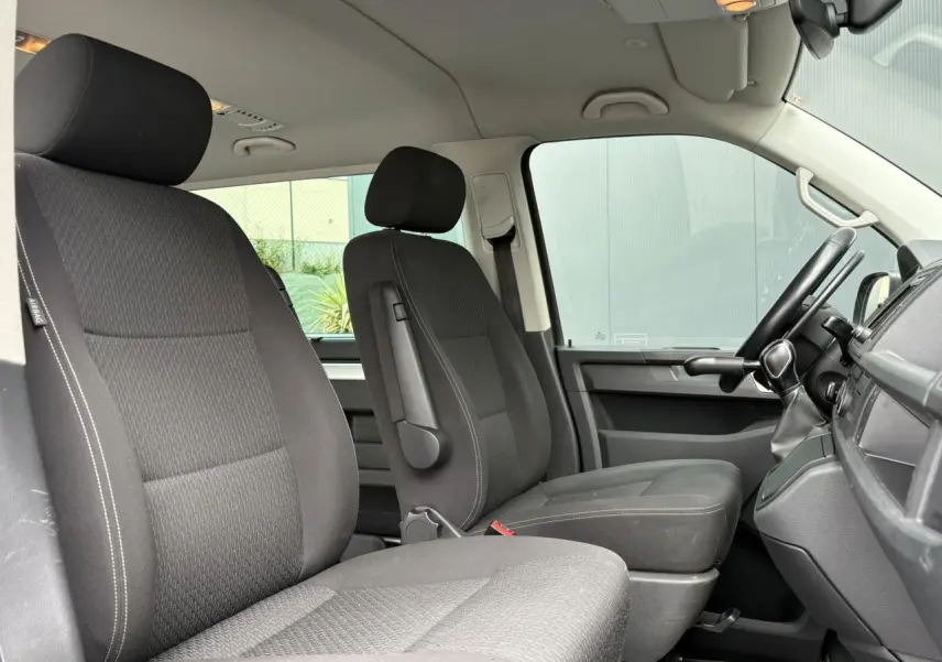 Vue intérieure côté conducteur du Volkswagen Multivan T6 gris Indium montrant les sièges avant en tissu noir et le tableau de bord.