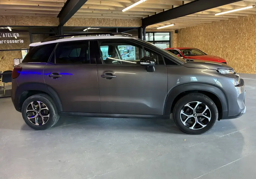 Profil droit d'un Citroën C3 Aircross gris platinium métal avec toit blanc et jantes alu bi-ton en intérieur d'atelier.