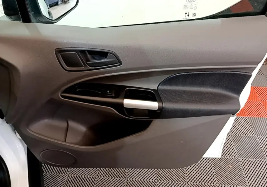 Vue intérieure côté gauche de la porte avant d’un Ford Transit Fourgon blanc, avec poignée argentée et panneau noir texturé.