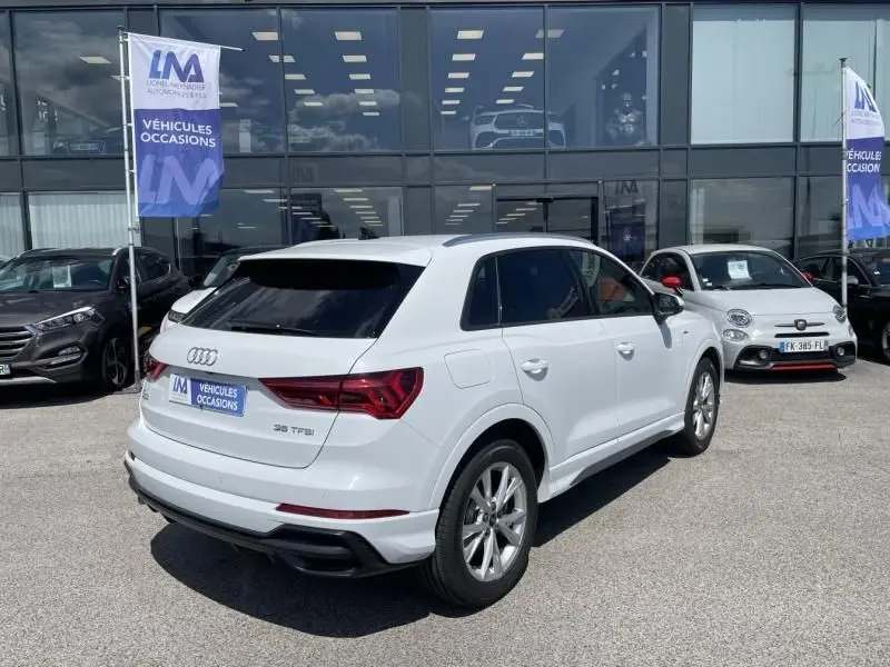 Audi Q3 blanc en 3/4 arrière droit, version S-Line avec détails noirs et vitres teintées, stationné devant un showroom.