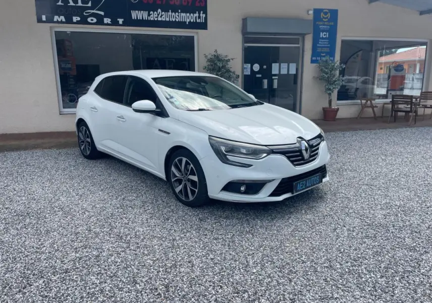Renault Mégane IV blanc vue 3/4 avant droit, avec jantes alliage et calandre chromée, stationnée sur gravier devant un commerce.