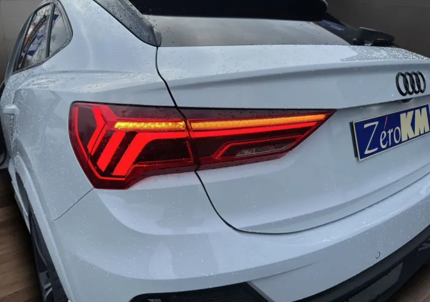 Vue 3/4 arrière droite d'un Audi Q3 Sportback blanc glacier avec feux arrière LED allumés et pluie sur la carrosserie.