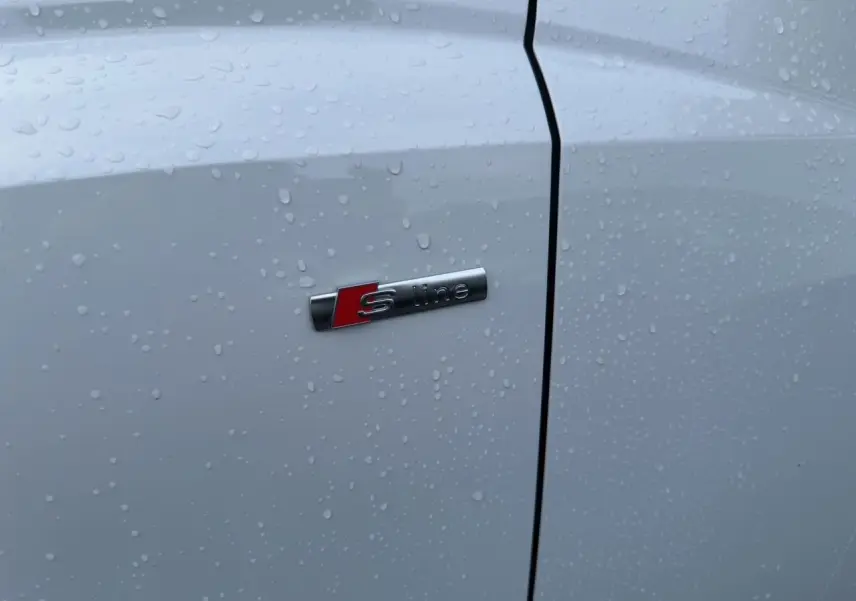 Gros plan sur l'emblème S line sur la porte blanche d'un Audi Q3 Sportback 2024 avec gouttes de pluie.