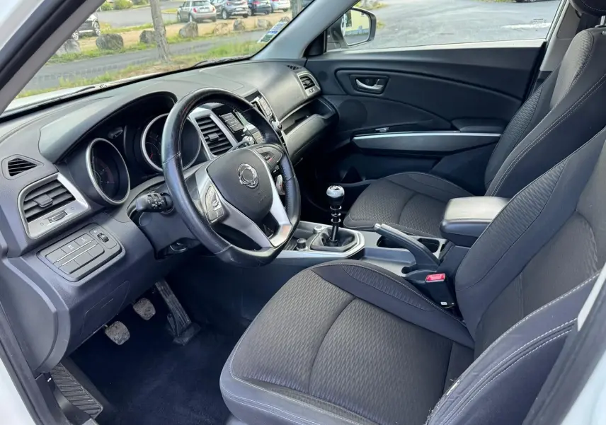 Intérieur côté conducteur du SsangYong Tivoli blanc 2016, volant cuir, levier de vitesse manuel et sièges tissu noirs.