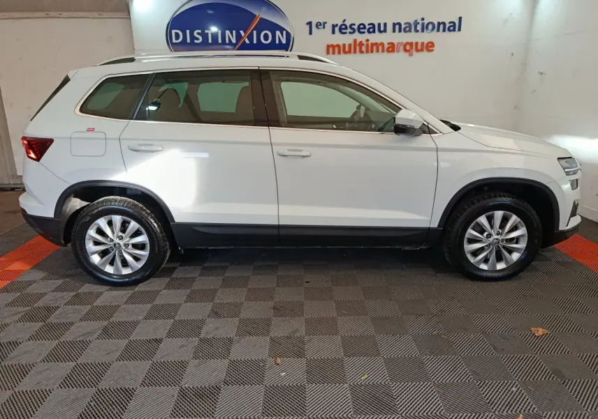 Vue latérale droite d'un Skoda Karoq blanc 2024 avec jantes alliage et barres de toit noires en intérieur showroom.