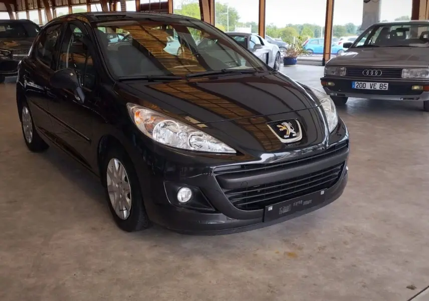 Peugeot 207+ noire vue 3/4 avant droit dans un garage, mettant en valeur ses phares et calandre distinctifs.