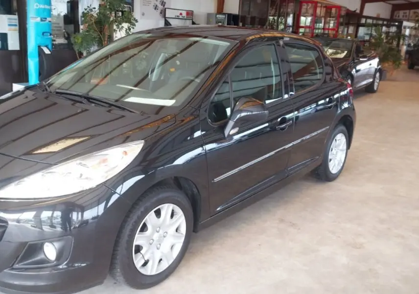 Peugeot 207+ noire vue en 3/4 avant droit dans un showroom, avec jantes argentées et phares allumés.