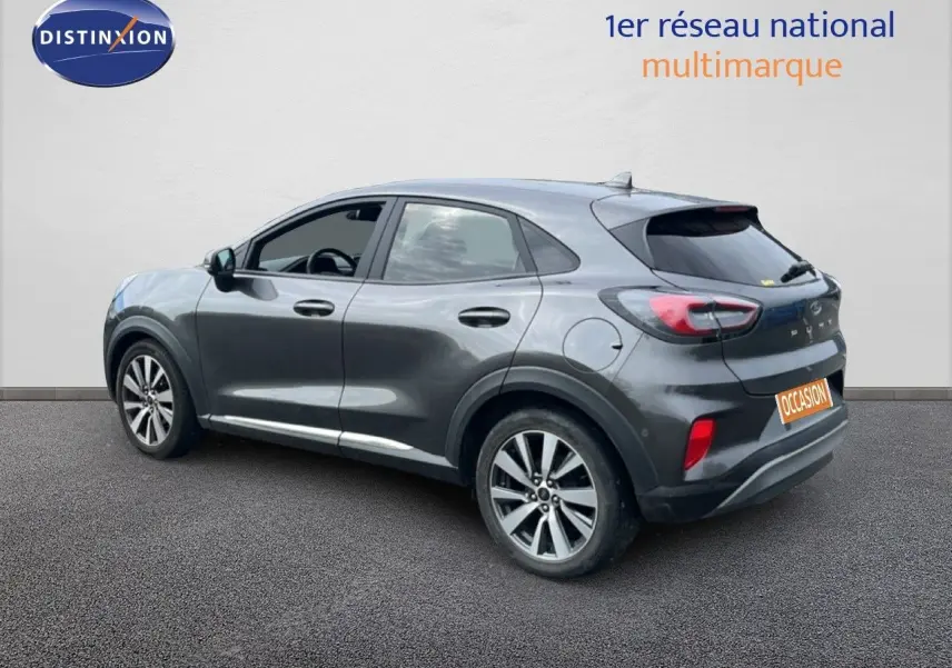 Ford Puma gris en vue 3/4 arrière droit, avec jantes alliage et vitres teintées, stationné devant un mur blanc.