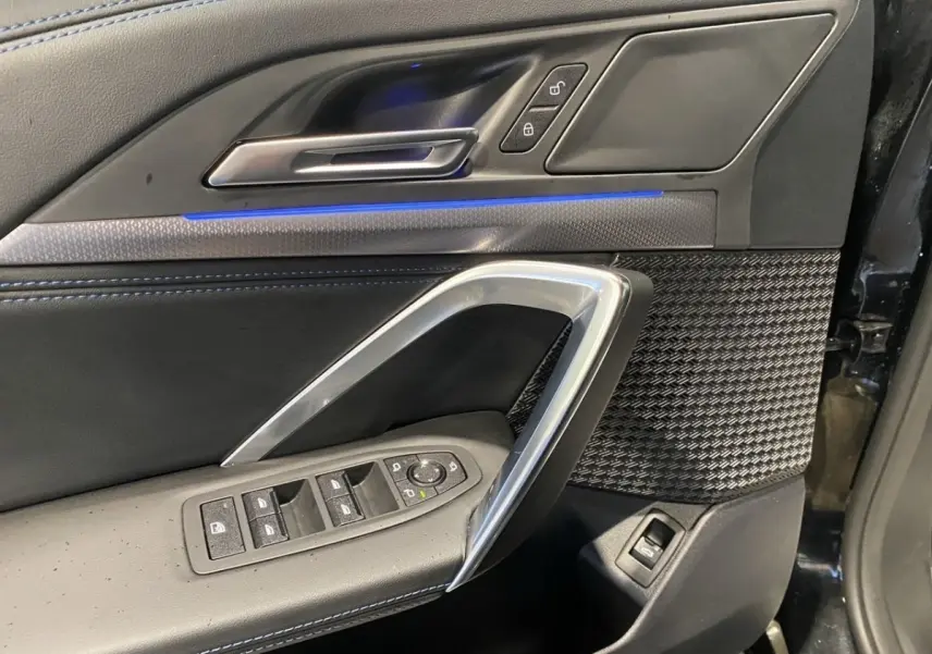 Vue rapprochée de la porte avant gauche noire du BMW X1 2025 avec éclairage d'ambiance bleu et commandes électriques.