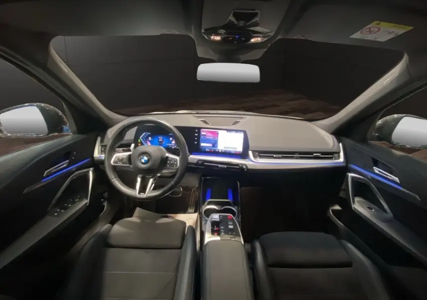 Intérieur noir du BMW X1 2025 vu de face, tableau de bord moderne avec écran tactile et éclairage d'ambiance bleu.