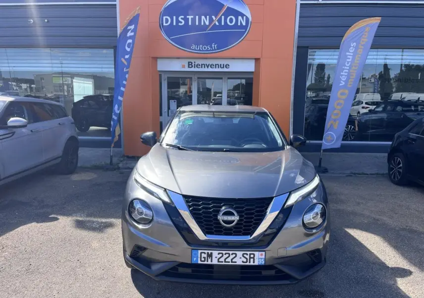 Nissan Juke 2023 gris argile vue de face devant une concession avec drapeaux publicitaires.