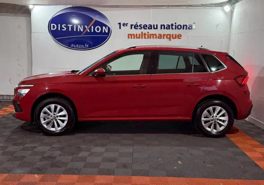 Vue latérale gauche d'un Skoda Kamiq rouge 2024 garé en intérieur avec jantes alliage et rails de toit noirs.