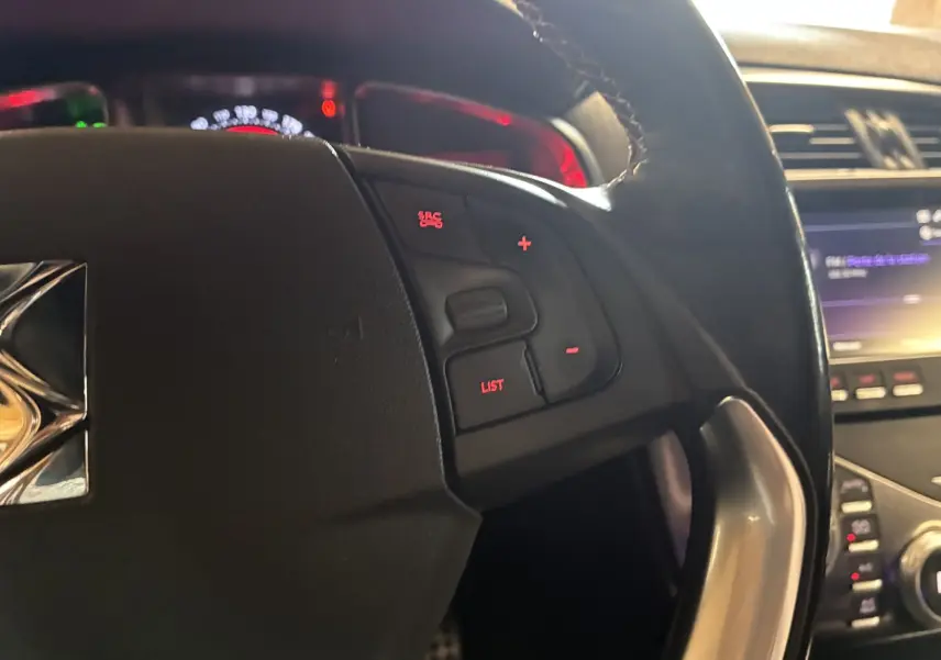 Gros plan sur le volant noir de la DS5 gris, avec commandes au volant éclairées en rouge et tableau de bord en arrière-plan.