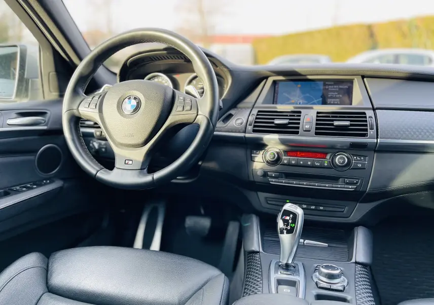 Vue intérieure du poste de conduite d'une BMW X6 blanche 50D M 2013, avec volant cuir M et écran central de navigation.