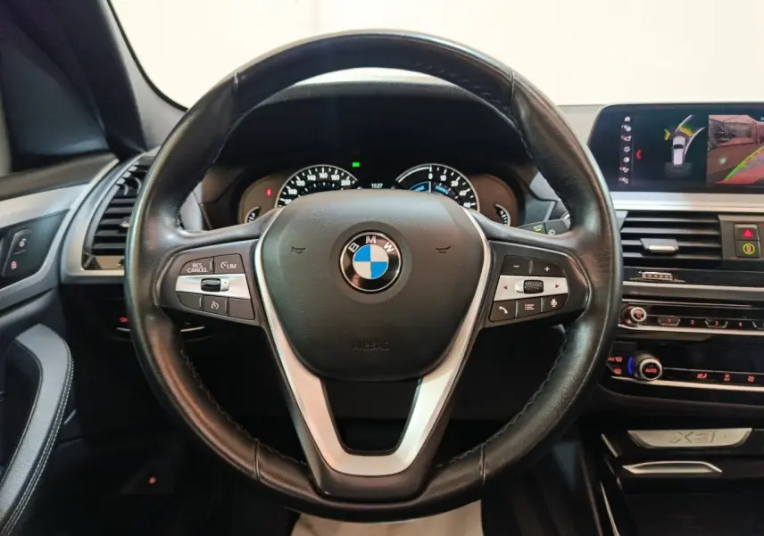 Vue intérieure centrée sur le volant cuir noir BMW X3 avec tableau de bord et écran multimédia en arrière-plan.