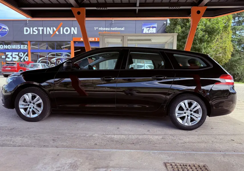 Profil côté gauche d'une Peugeot 308 SW noire métal 2018 sous abri avec jantes alliage argentées.