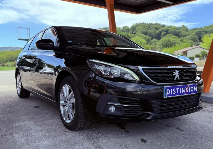 Peugeot 308 SW noir métal vue 3/4 avant droit sous un abri avec plaque Distinxion visible.