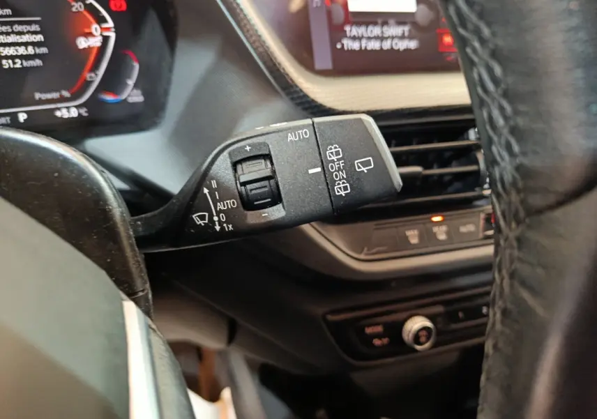 Gros plan sur la commande d’essuie-glace à gauche du volant d’une BMW Série 1 noire, avec tableau de bord numérique en arrière-plan.