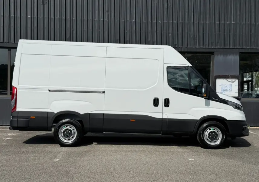 Vue de profil côté droit d’un utilitaire Iveco Daily blanc avec bas de caisse noir, stationné devant un mur sombre.