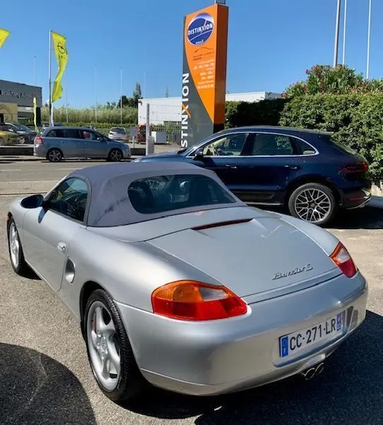 Vue 3/4 arrière d'une Porsche Boxster 986 gris argent avec capote noire, stationnée en extérieur par temps clair.