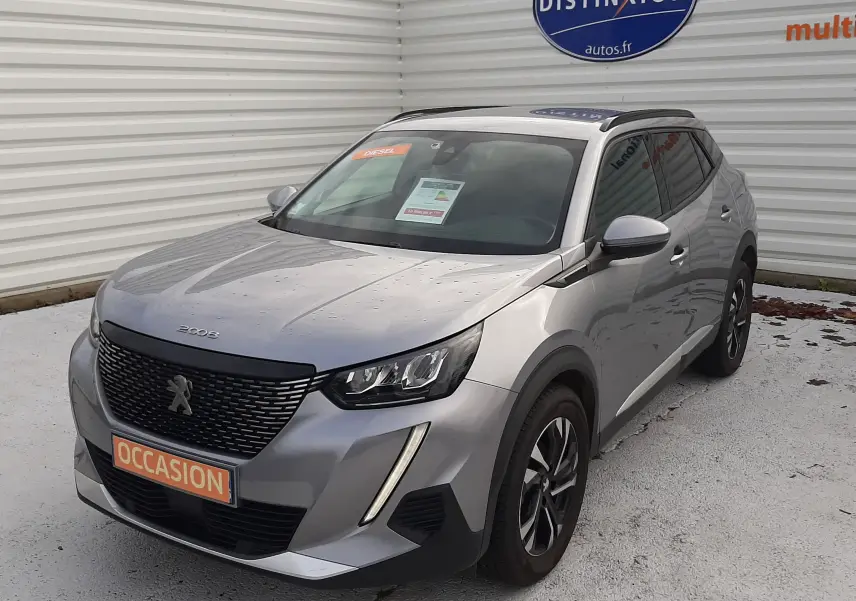 Peugeot 2008 gris en 3/4 avant droit, avec calandre noire et feux LED, stationné devant un mur blanc.