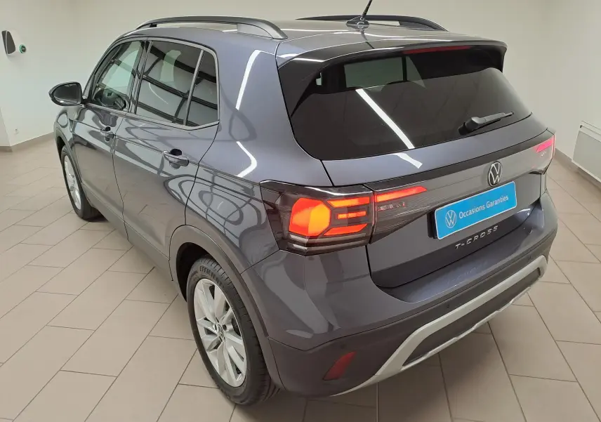 Volkswagen T-Cross gris cendré en 3/4 arrière droit, feux arrière allumés et toit avec barres de toit.