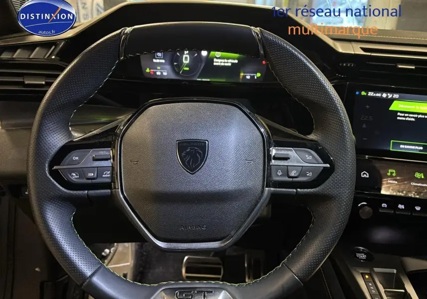 Vue rapprochée du volant cuir noir perforé avec logo Peugeot et badge GT, tableau de bord numérique en arrière-plan.