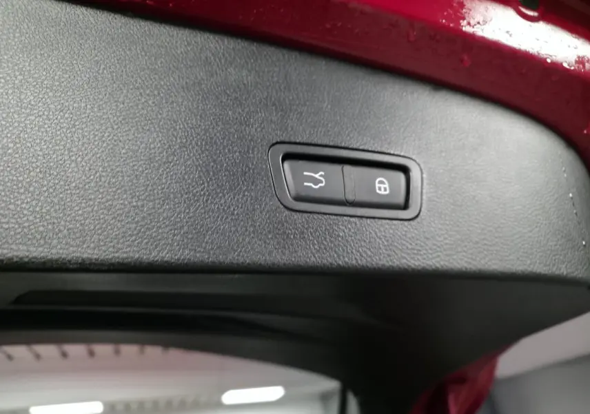 Gros plan sur le bouton de fermeture électrique du hayon, intérieur noir, bord rouge du coffre visible.