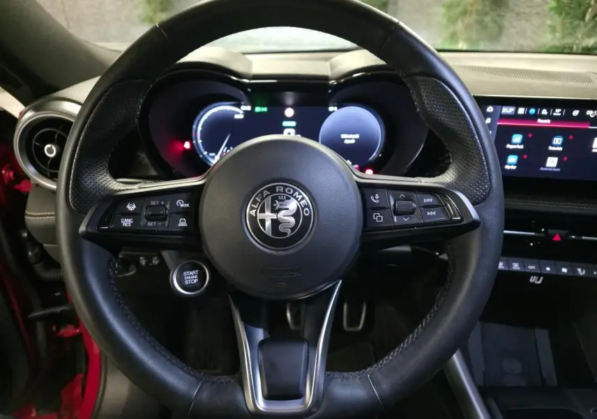Vue rapprochée du volant sport en cuir noir de l'Alfa Romeo Tonale rouge, avec tableau de bord digital et bouton start visible.