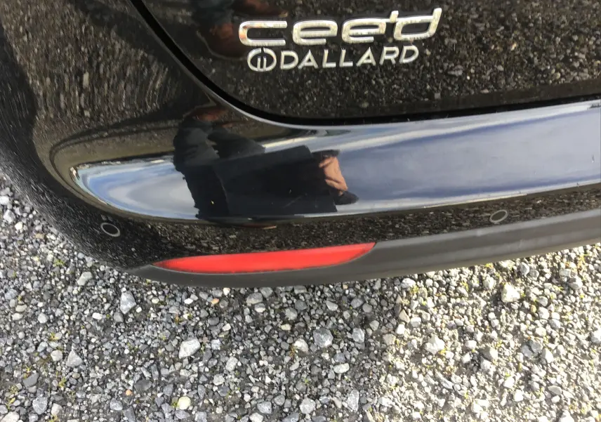 Gros plan sur le logo arrière et le pare-chocs noir brillant d'une KIA CEE'D SW 2017, avec reflet et sol gravillonné.