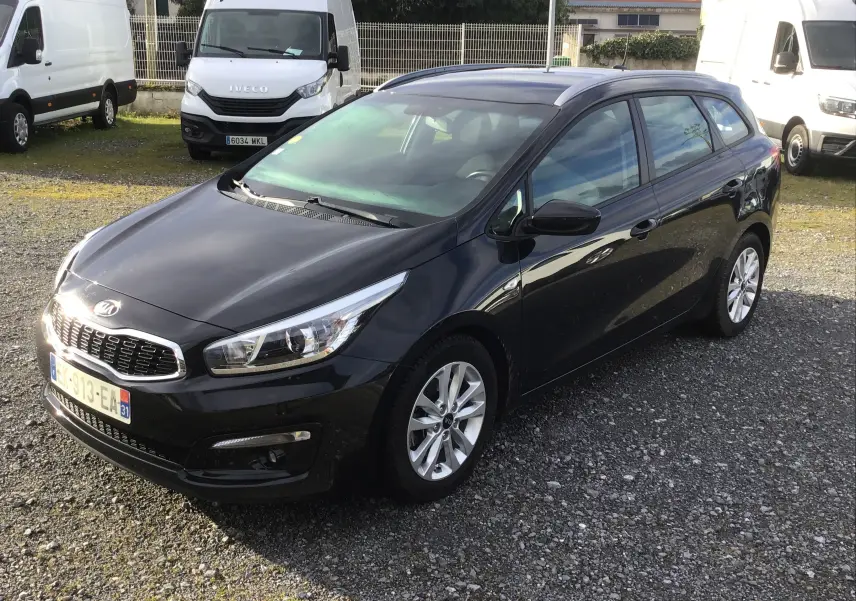 KIA CEE'D SW noir vue 3/4 avant droit, stationnée sur un sol gravillonné avec des fourgons blancs en arrière-plan.