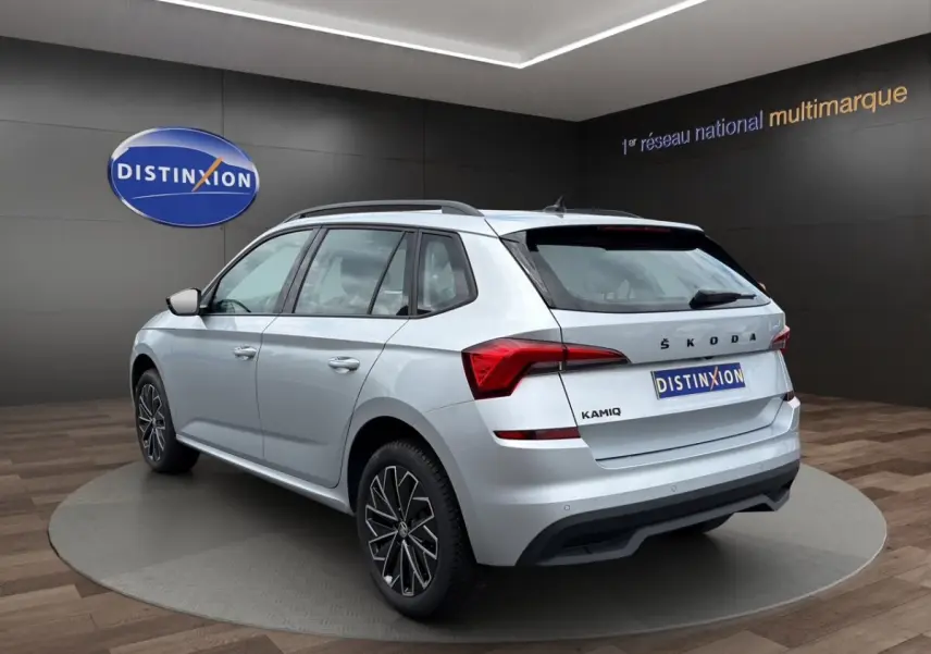 Vue 3/4 arrière droite d'un Skoda Kamiq gris clair avec jantes bi-ton et barres de toit noires en showroom.