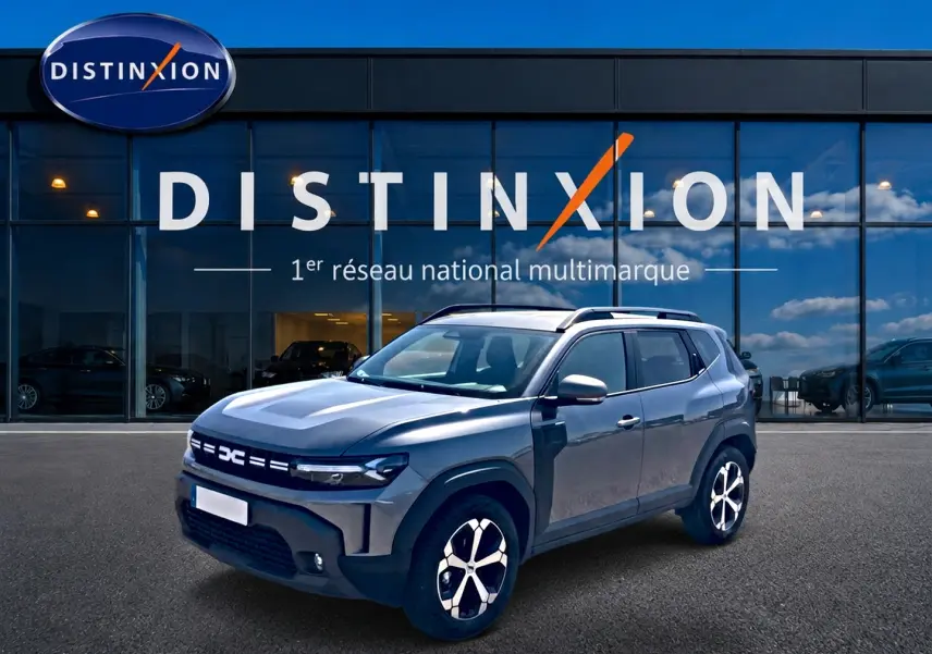 Vue 3/4 avant droit du Dacia Duster gris schiste avec jantes alu et barres de toit devant une vitrine Distinxion.