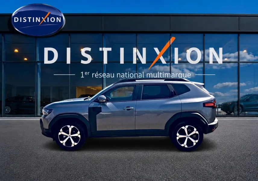 Profil droit du Dacia Duster gris schiste 2026 avec jantes alu et barres de toit visibles devant une vitrine Distinxion.