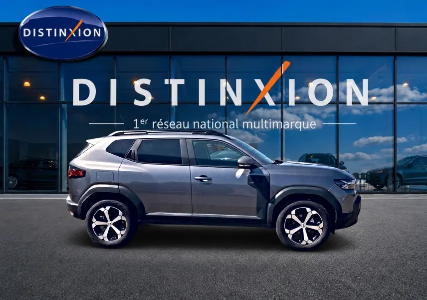 Vue de profil côté gauche d'un Dacia Duster gris schiste 2026 avec jantes alu et barres de toit, devant une vitrine Distinxion.