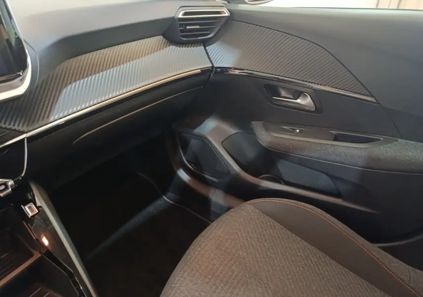 Vue intérieure côté passager de la Peugeot 208 Hybrid 2025, tableau de bord avec finition carbone et siège tissu gris.