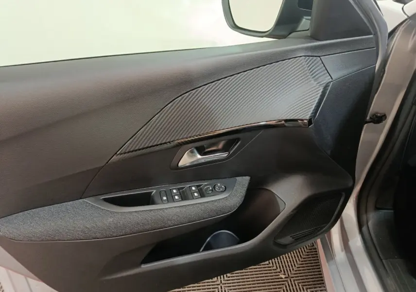 Vue intérieure côté conducteur de la porte grise claire d'une Peugeot 208 Hybrid 2025 avec habillage carbone et commandes électriques.