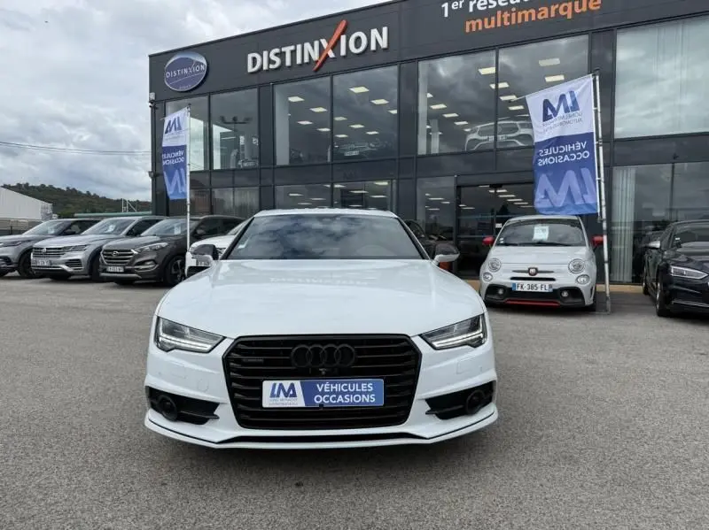 Vue frontale d'une Audi A7 Sportback blanche 2018 avec calandre noire et plaque « VÉHICULES OCCASIONS » devant un showroom.