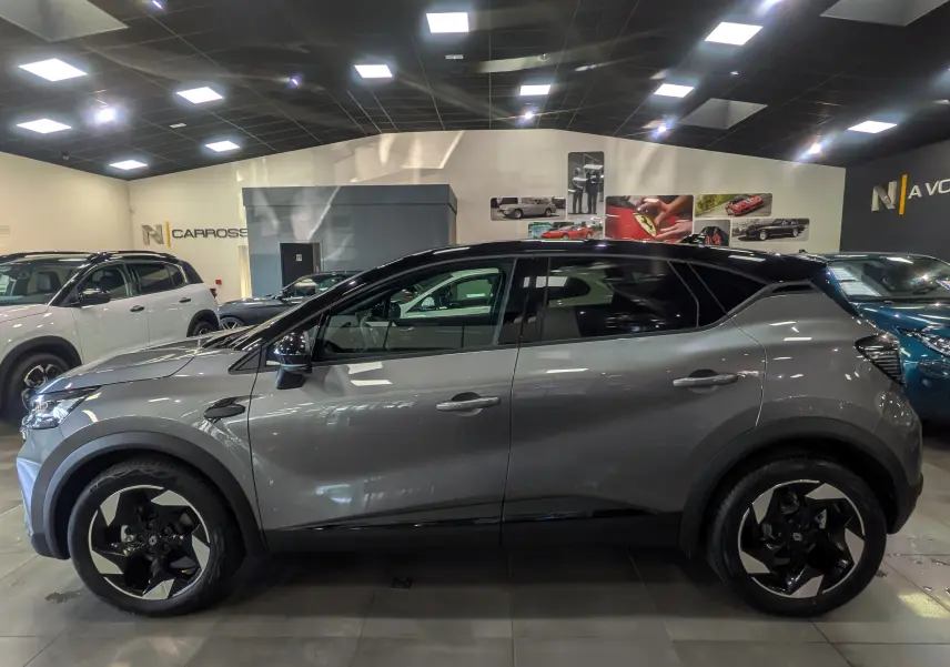 Profil côté gauche du Renault Captur 2025 gris Cassiopé avec toit noir et jantes biton dans un showroom.