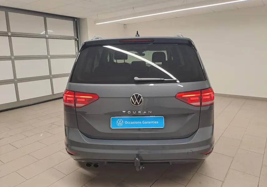 Vue arrière d'un Volkswagen Touran gris dauphin 2024 avec attelage et feux allumés en intérieur.