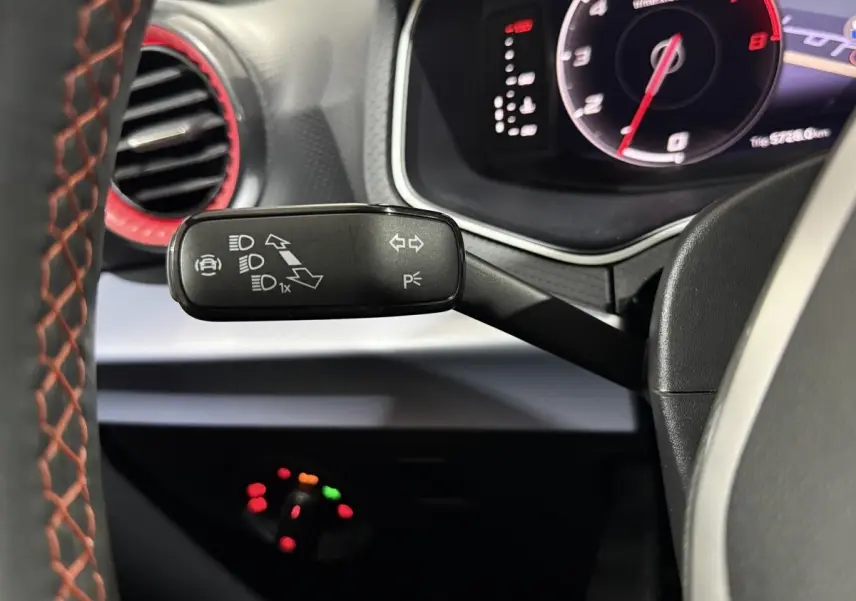 Gros plan sur la manette de clignotants noire avec icônes blanches, volant cuir gris foncé avec surpiqûres rouges, tableau de bord digital.